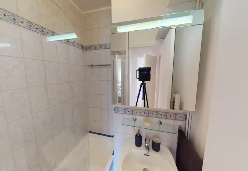 Compact tiled bathroom with a bathtub, wall-mounted mirror cabinet and a sink with accessories. Neutral light and decorative blue tile border create a clean, functional space.