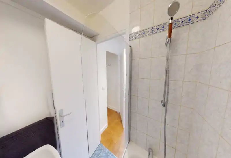 Compact bathroom with tiled shower corner and a handheld showerhead. The image shows the shower area, tiled walls and part of the doorway leading to a wooden-floored hallway.