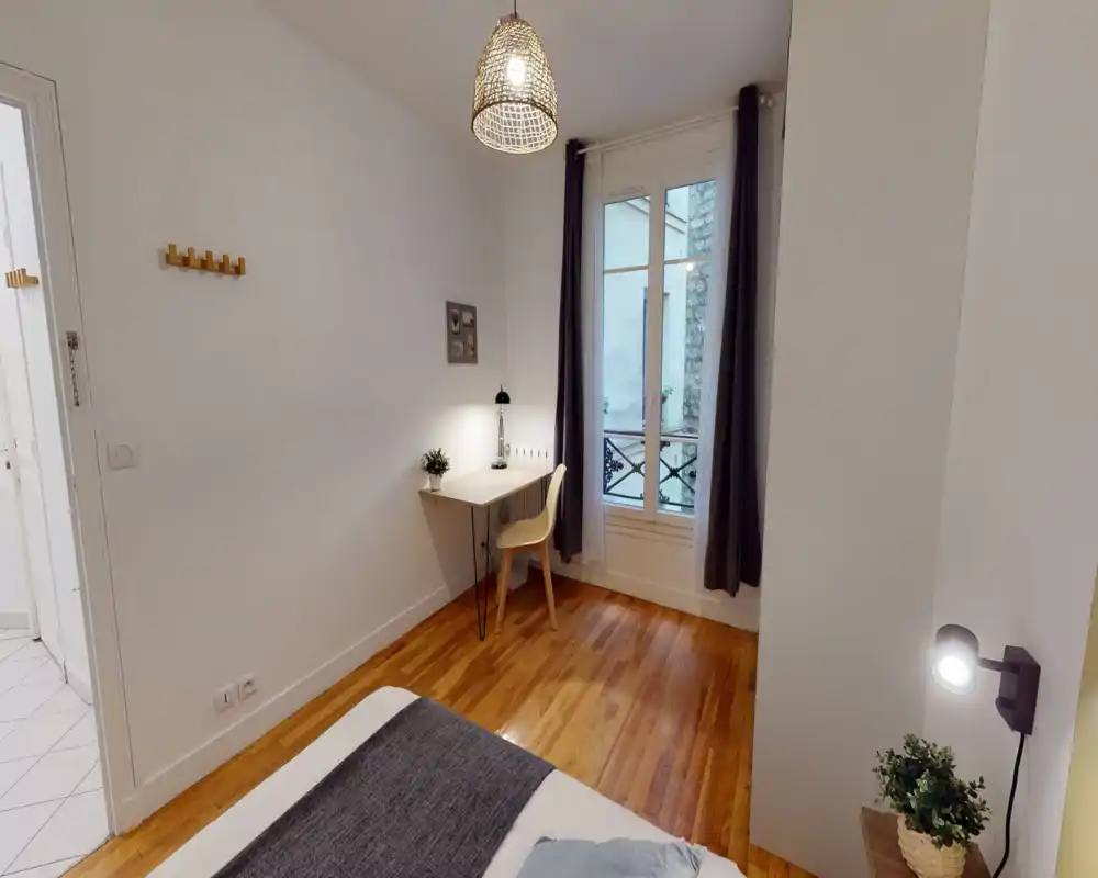 Small, bright bedroom with wooden floors, a visible double bed in the foreground, a compact desk by the window and soft, modern lighting—ideal for a single or couple seeking a cozy city room.