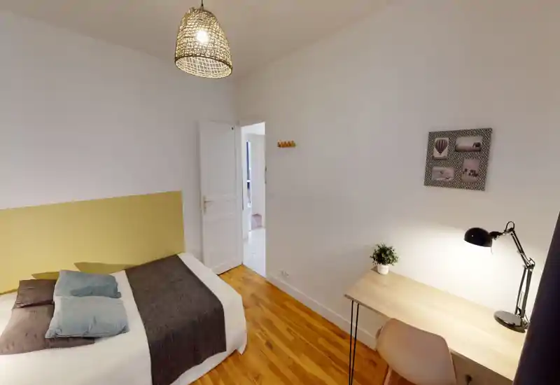Bright, neatly staged bedroom with a double bed, soft pillows and a cozy throw; a wooden desk with a lamp offers a functional workspace.