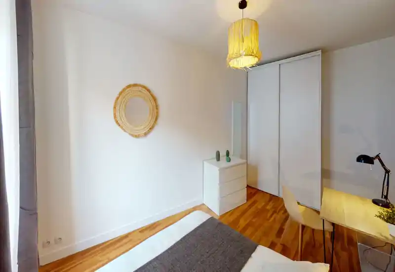 Chambre lumineuse et épurée avec lit visible au premier plan, murs blancs, parquet, grand placard coulissant et petit bureau avec lampe — ambiance chaleureuse et accueillante.