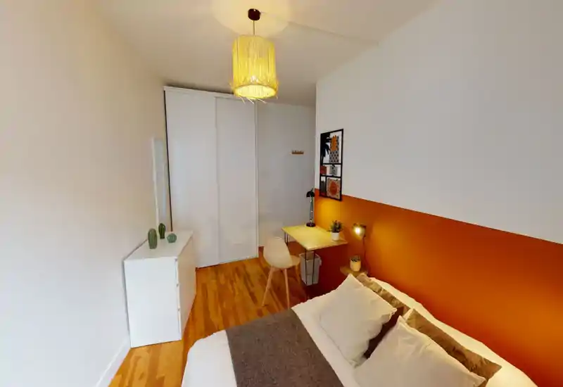 Chambre lumineuse et décorée avec un mur d'accent orange, lit confortable et un petit espace bureau — idéale pour une personne ou un invité. Sol en bois, placard coulissant et suspension lumineuse chaleureuse.