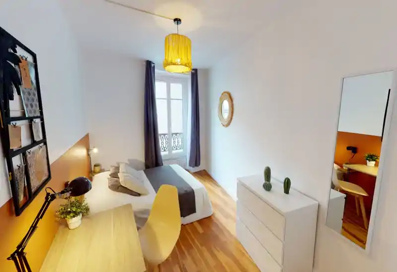 Chambre lumineuse et décorée avec un lit double, sol en bois et un bureau près du lit. La pièce présente des accents chaleureux, une grande fenêtre avec rideaux et une glace pleine longueur, idéale pour une annonce locative.