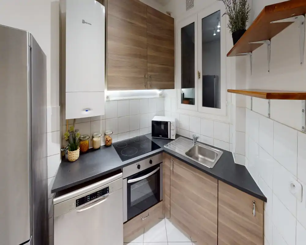 Cuisine compacte et moderne avec meubles effet bois, plaque électrique et four encastré. Évier en acier inoxydable, lave-vaisselle et réfrigérateur pour un espace fonctionnel et bien organisé.