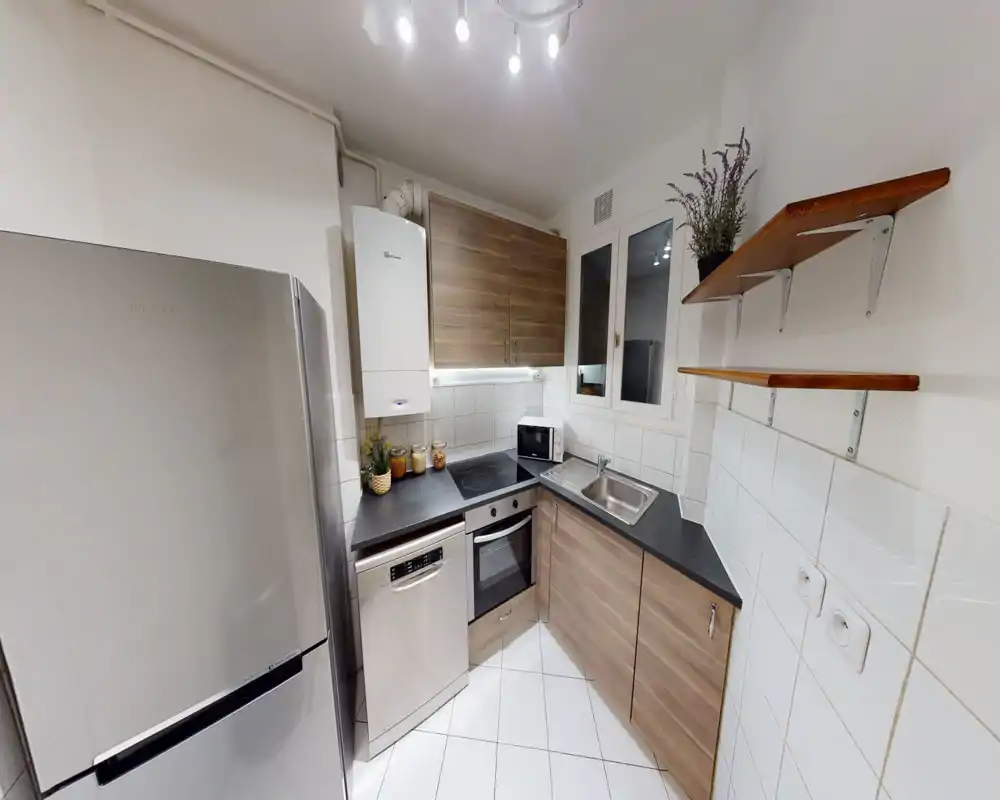 Cuisine moderne compacte avec placards en bois, plan de travail noir, four et lave-vaisselle encastrés et réfrigérateur en inox. Espace lumineux et soigné avec étagères ouvertes et petites plantes décoratives.