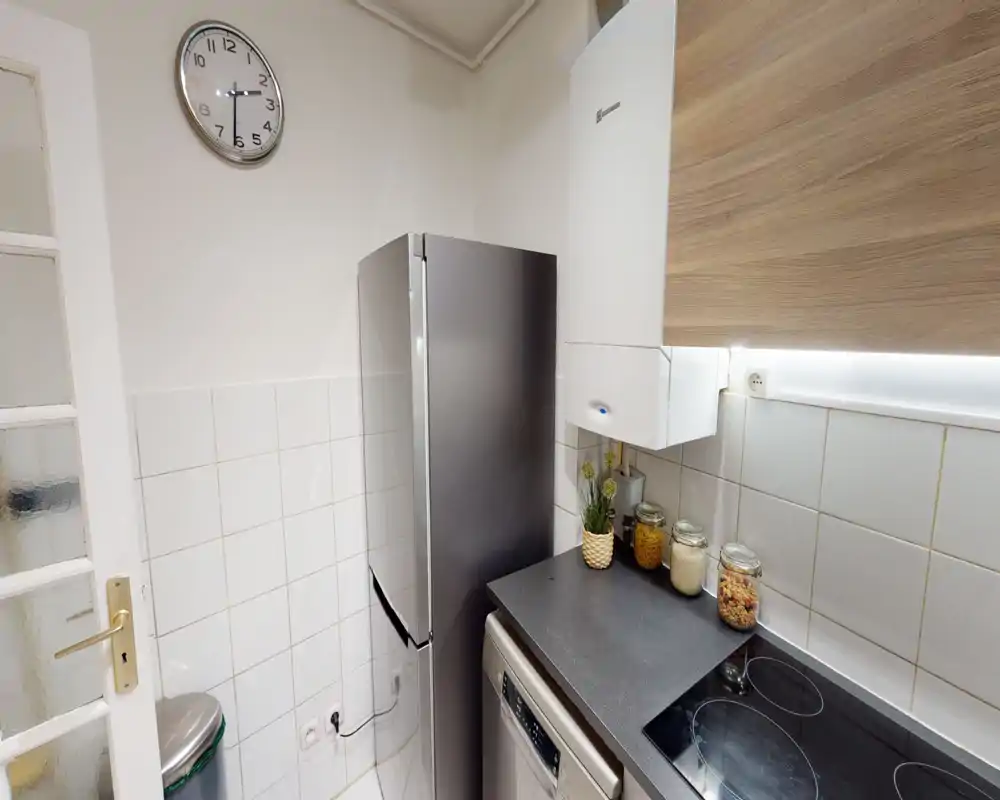 Coin cuisine moderne et compact avec un grand réfrigérateur inox, plaque à induction, lave-vaisselle et bocaux sur le plan de travail; carrelage blanc et meuble bois pour un espace propre et fonctionnel.