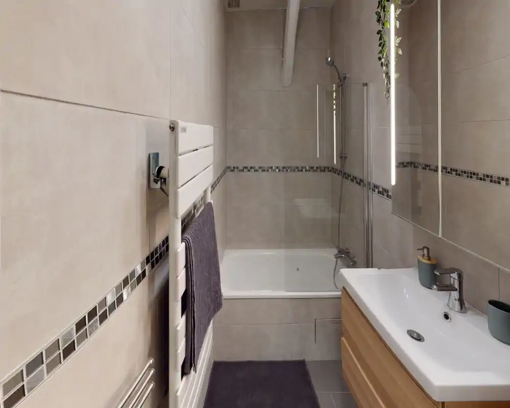 Salle de bain moderne carrelée avec baignoire et paroi de douche, lavabo mural avec meuble en bois, radiateur porte-serviettes et miroir ; tons neutres et finitions soignées.