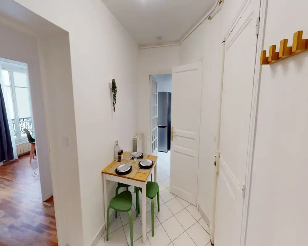 Coin cuisine/salle à manger avec petite table en bois pour deux, tabourets verts et sol carrelé ; espace lumineux, simple et fonctionnel, idéal pour un appartement en ville.