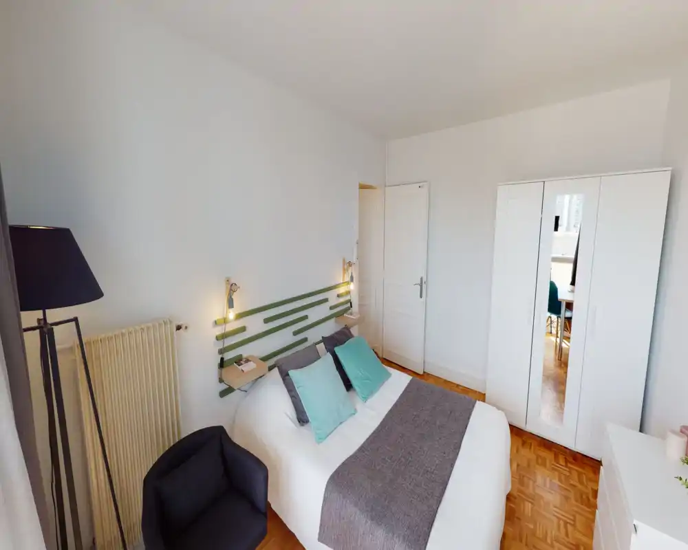 Bright, modern bedroom with a double bed, soft textiles and a mirrored wardrobe. The room features a decorative headboard, bedside shelves with lamps, a comfortable armchair and warm parquet flooring.
