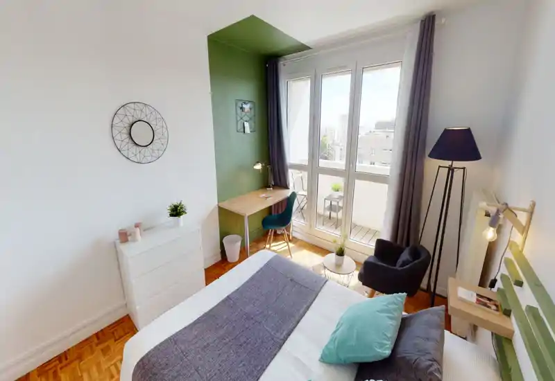 Bright, modern bedroom with a large window and balcony access, a comfortable double bed dressed with pillows and throw, bedside lamp and a cozy reading chair. The small desk in the green alcove creates a practical work corner while the parquet floor and decorative accents add warmth.