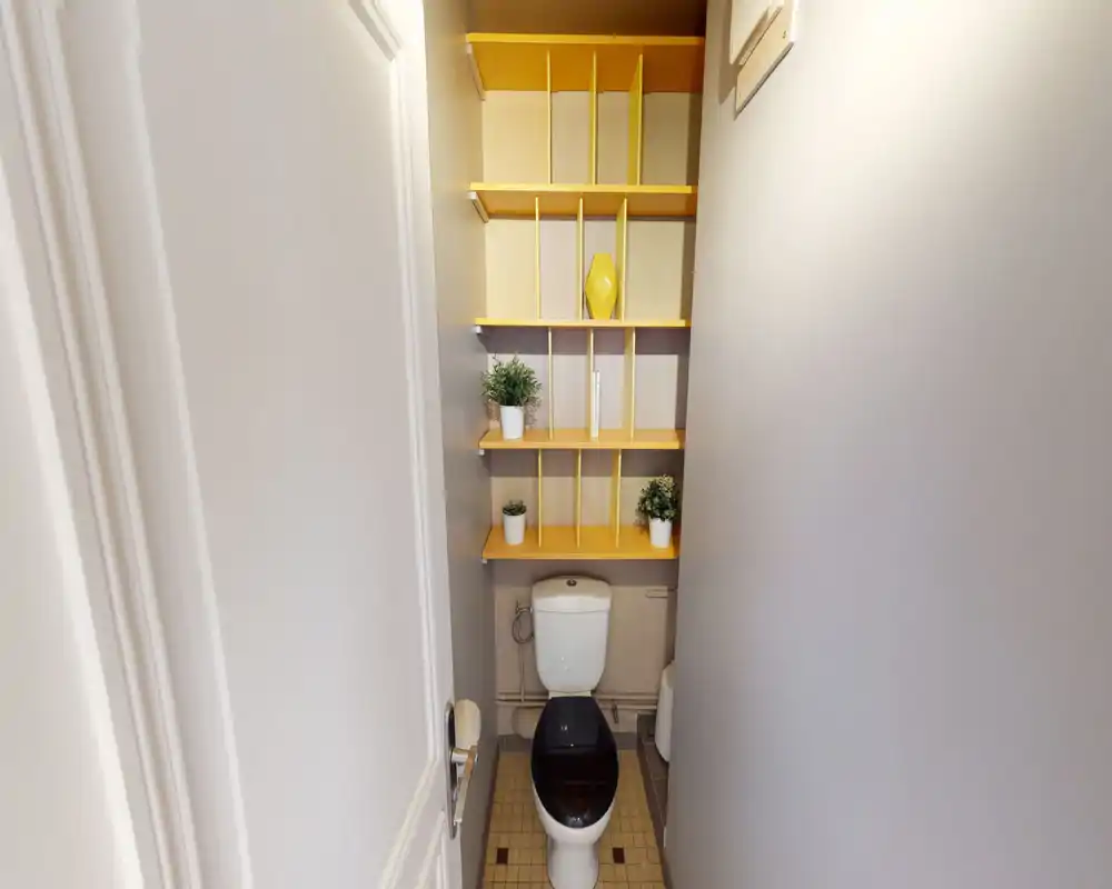 Compact bathroom with a toilet beneath built-in yellow shelving and decorative plants; clean and well-lit small WC.