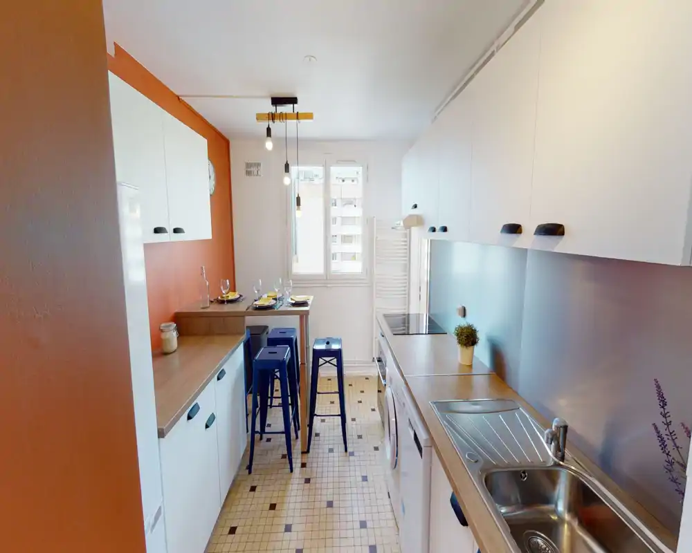 Bright, compact kitchen with modern white cabinets, wooden countertops and a tiled floor. A small breakfast bar with blue stools and decorative details (plant, tableware) make the space inviting and functional.