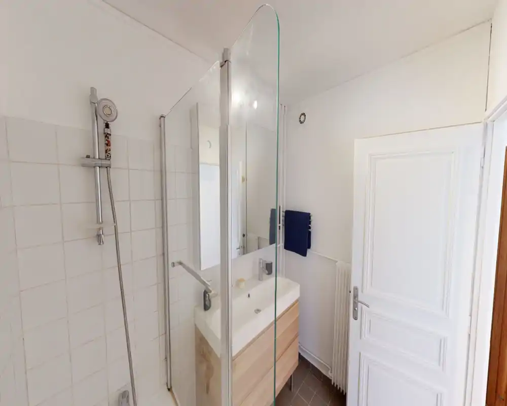 Bright, modern bathroom with a walk-in shower, glass partition and a vanity with sink and mirror. Clean white tiles and wooden-tone cabinet create a fresh, minimal look.