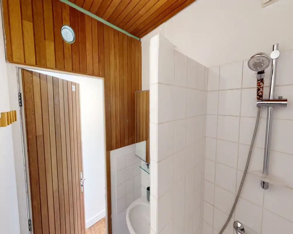 Compact bathroom with a tiled shower and handheld chrome showerhead; a small sink and wooden door/ceiling details add warmth.