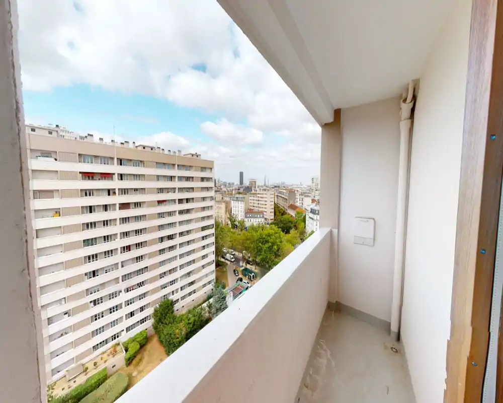 Small covered balcony with a city view; concrete floor and low parapet offer an outdoor space overlooking neighboring buildings and treetops.