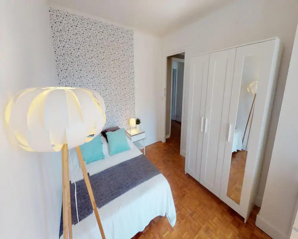 Camera da letto luminosa e ben arredata con letto matrimoniale in lenzuola bianche, cuscini turchesi e comodino con lampada. Un pannello di carta da parati decorativa e un grande armadio bianco con specchio completano lo spazio moderno e arioso.