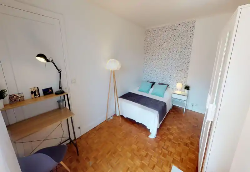 Camera da letto luminosa e ordinata con letto matrimoniale, parete decorativa e comodino; pavimento in legno e una piccola scrivania rendono l'ambiente funzionale e accogliente.