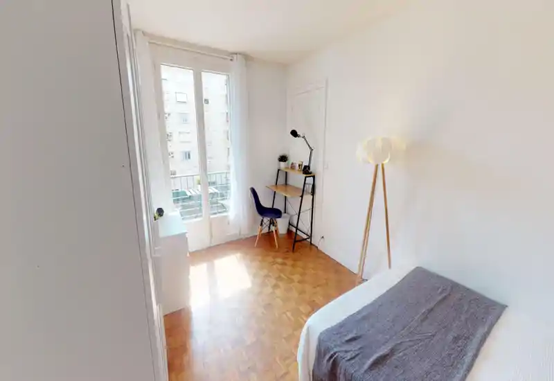Camera da letto luminosa e minimale con pavimento in parquet, letto ben rifatto in primo piano, lampada da terra su treppiede e una piccola scrivania davanti a una grande finestra che lascia entrare molta luce naturale.