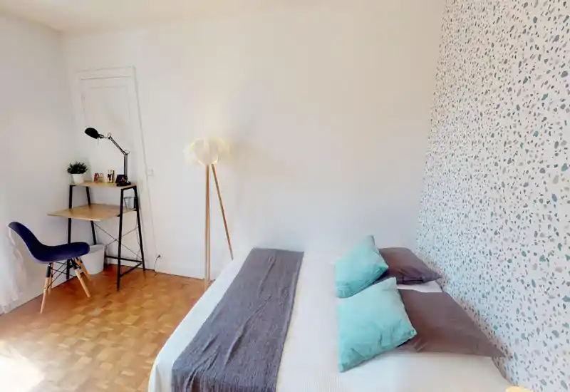 Camera da letto luminosa e moderna con un letto matrimoniale ben rifatto, cuscini decorativi e una parete accentata a motivo. Una piccola scrivania e una sedia vicino alla finestra rendono la stanza adatta anche per lavorare.