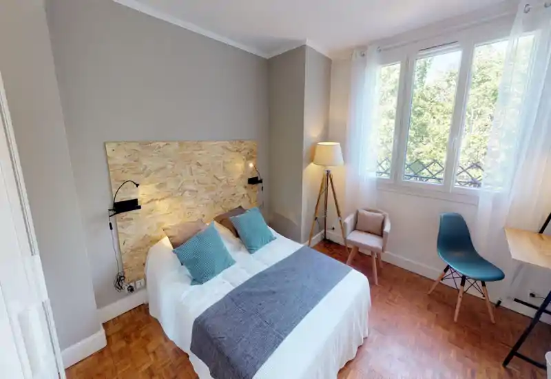 Chambre lumineuse et soignée avec lit double, tête de lit en panneaux de bois et grande fenêtre apportant une belle lumière naturelle.