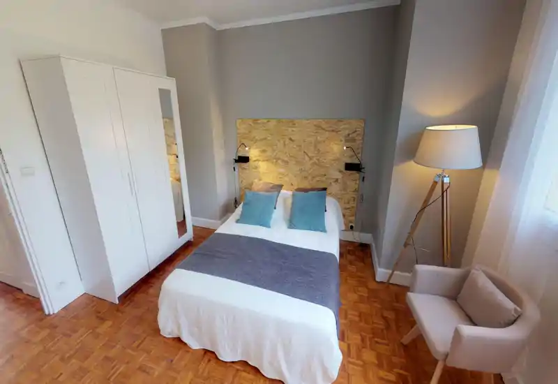 Chambre claire et stylée avec lit central devant une tête de lit en bois texturé, lampes de lecture, armoire avec miroir et un fauteuil confortable près de la fenêtre.