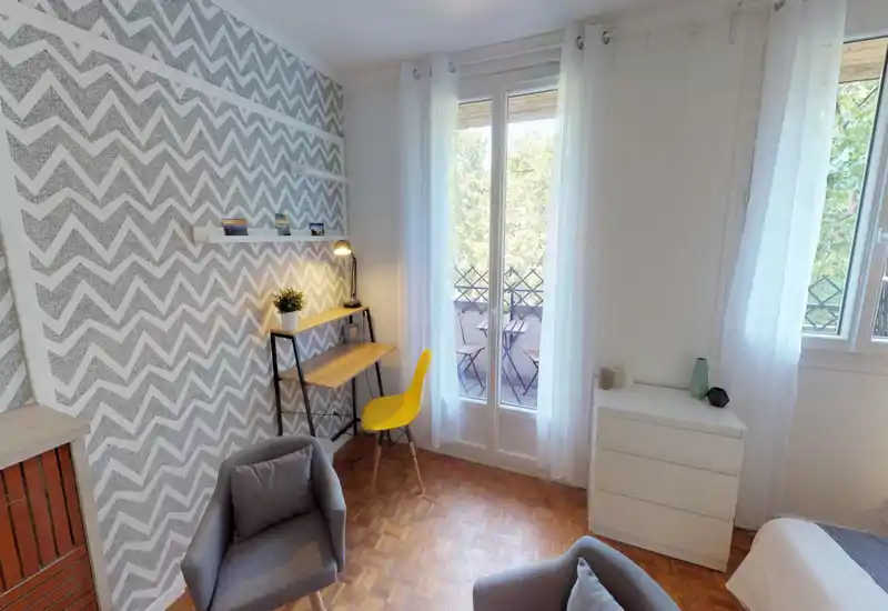 Angolo camera da letto luminoso e accogliente con parete a motivo geometrico, una scrivania e balcone. Sono visibili poltrone, cassettiera e parte del letto, adatto per un soggiorno confortevole.