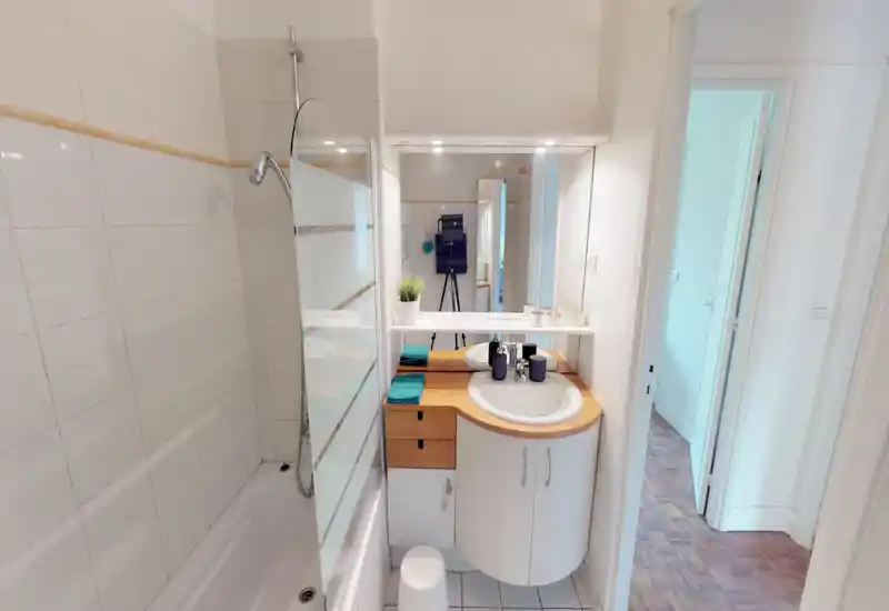 Bagno compatto e luminoso con vasca/doccia, paratia in vetro e mobile lavabo curvo con specchio. Piastrelle bianche e piano in legno donano un aspetto pulito e ordinato.