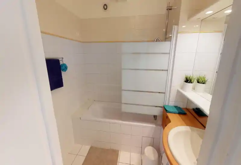Bagno piastrellato luminoso con vasca e parziale pannello di vetro per la doccia, lavabo bianco su mobile in legno e piccole piante decorative sul ripiano dello specchio.
