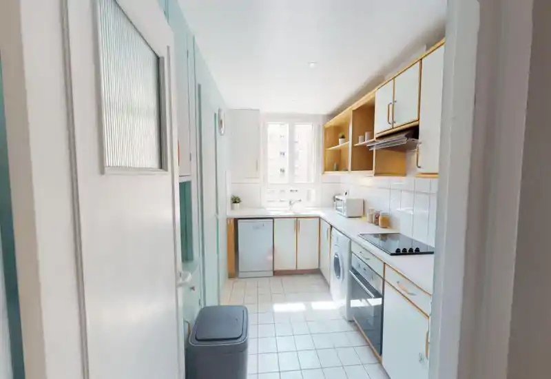 Cucina compatta e luminosa con mobili bianchi, elettrodomestici integrati (forno, piano cottura, lavastoviglie, lavatrice) e una finestra ampia che garantisce luce naturale. Pavimento piastrellato e scaffalature aperte per un ambiente ordinato e funzionale, ideale per annunci.