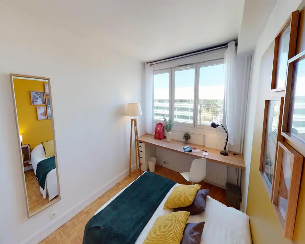 Chambre lumineuse et confortable avec lit bien fait, coussins d'accent et un large bureau sous une grande fenêtre — idéale pour se détendre et travailler depuis chez soi.