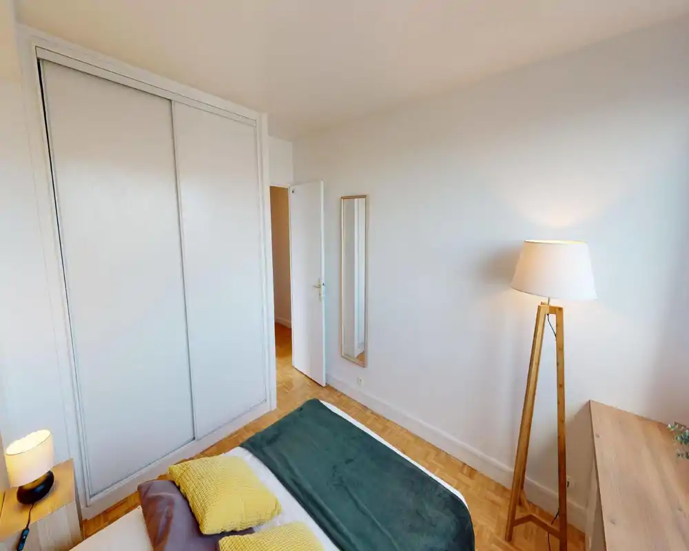 Chambre claire et soignée avec lit double habillé de textiles verts et jaunes, parquet, placard coulissant intégré et lampe sur pied chaleureuse — idéale pour une annonce.