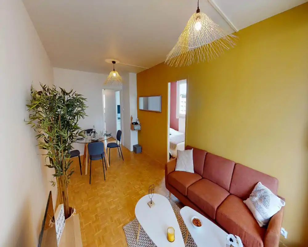 Salon lumineux et moderne avec un mur d'accent moutarde, un canapé couleur rouille, tables basses et une zone repas en arrière-plan. Suspensions contemporaines et parquet en bois pour une ambiance chaleureuse.