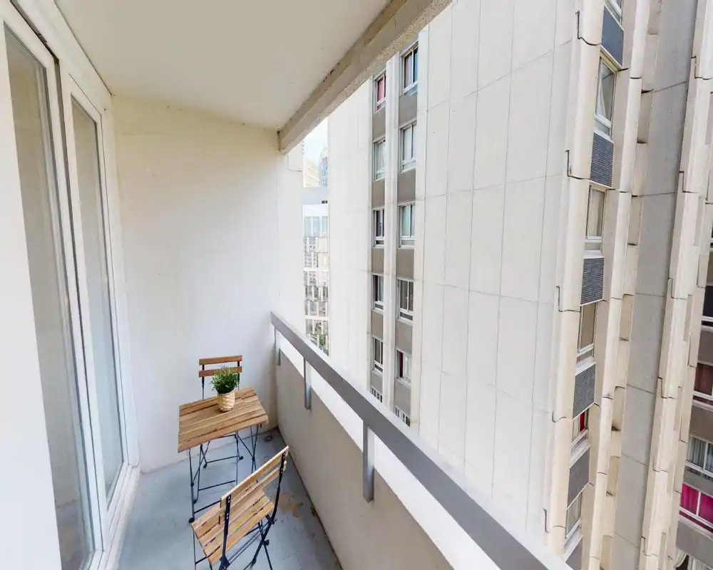 Petit balcon couvert avec une table bistro en bois et deux chaises pliantes, une plante en pot et vue sur des immeubles voisins.