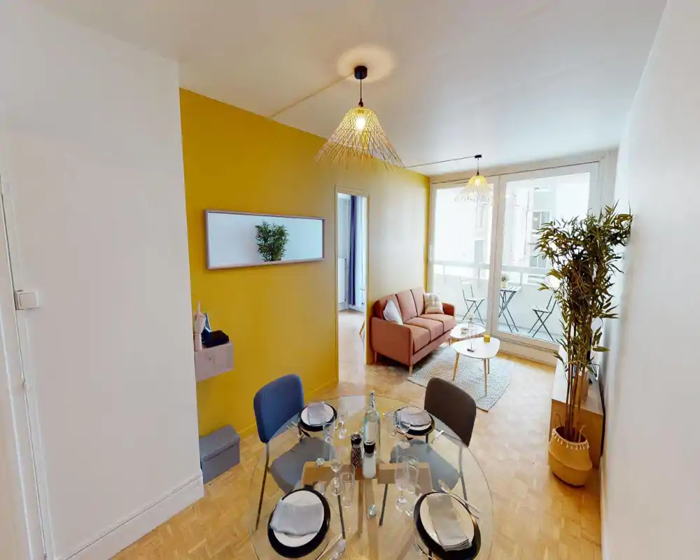 Espace de vie lumineux et bien mis en scène avec une petite table à manger au premier plan, un canapé confortable et de grandes baies vitrées donnant sur un balcon — un mur jaune et des plantes apportent une ambiance chaleureuse.