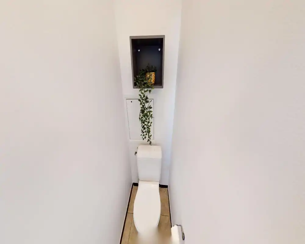 Toilettes compactes et bien éclairées avec WC blanc, sol carrelé et petite étagère murale avec plante retombante en décoration.