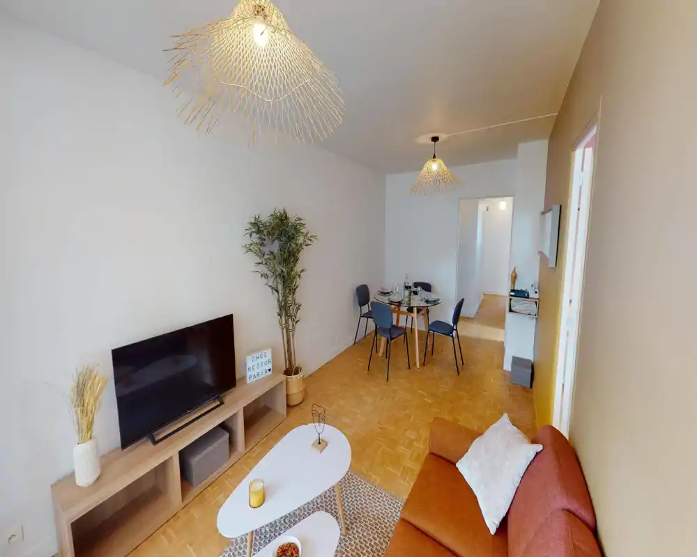 Salon spacieux et lumineux avec canapé couleur rouille, meuble TV et coin repas en arrière-plan. Parquet en bois naturel et éclairage chaleureux créent une ambiance accueillante.