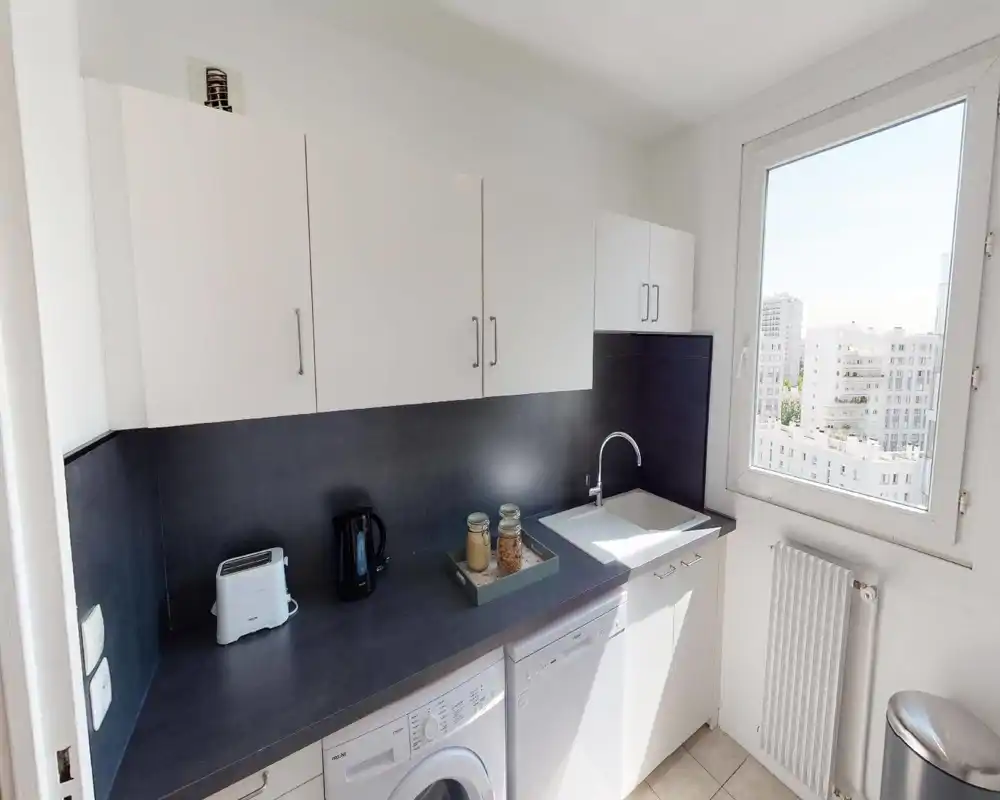 Cuisine moderne et compacte, lumineuse, avec placards blancs, plan de travail foncé, évier intégré et appareils électroménagers; grande fenêtre offrant une vue sur la ville.
