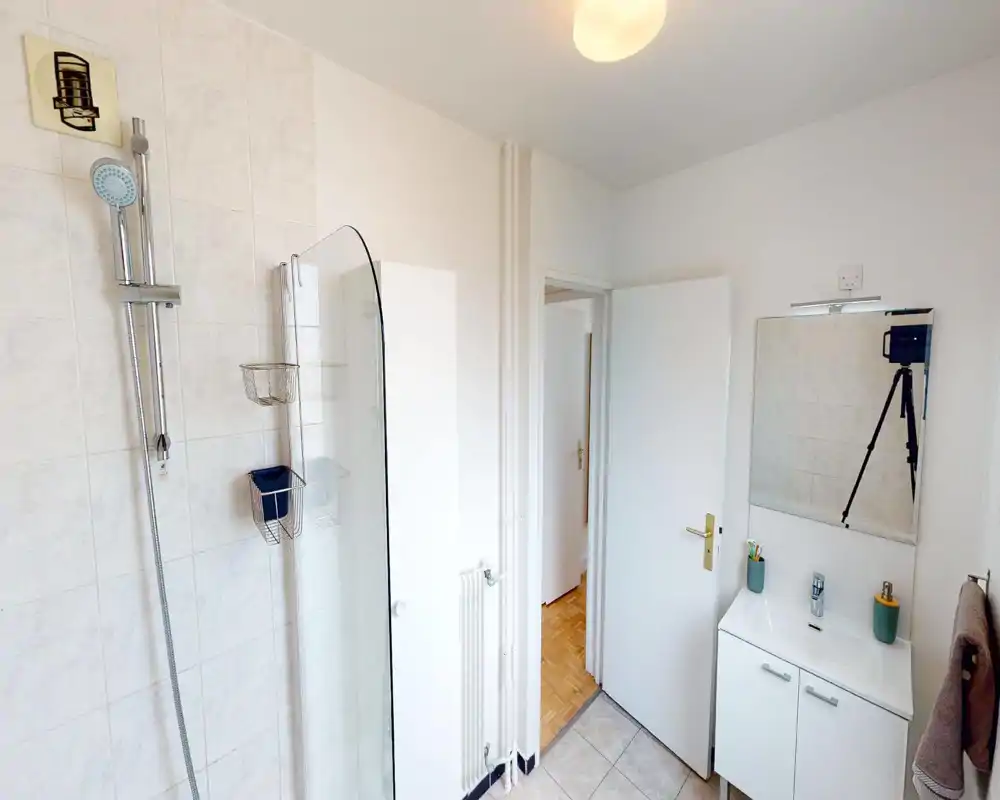 Salle de bain lumineuse et propre avec douche carrelée, paroi en verre, meuble vasque et miroir ; tons neutres et équipements simples pour un espace fonctionnel.