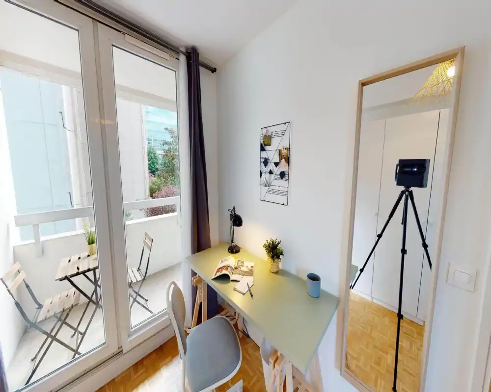 Bright, compact living area with a small desk and chair next to large sliding doors that open onto a balcony with bistro seating. The space features a desk lamp, decorative plant and a full-length mirror, ideal for a home office corner within a living room.
