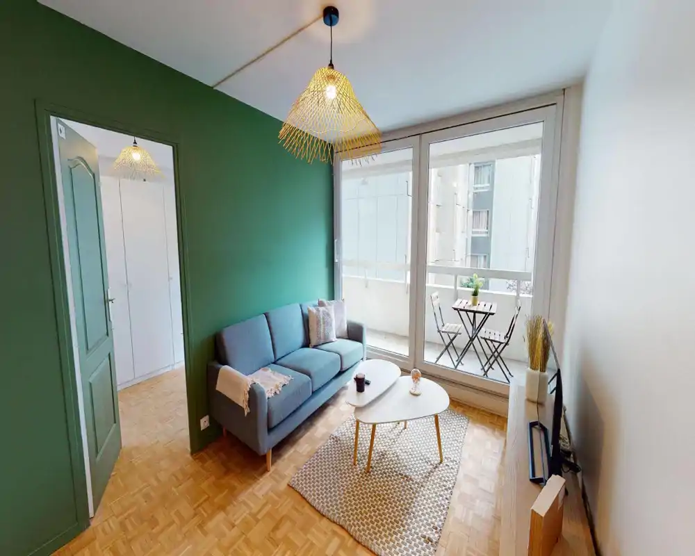 Bright, stylish living room with a comfortable three-seat sofa, coffee tables and a rug; large sliding windows open to a small balcony with bistro seating — ideal for relaxing and natural light.