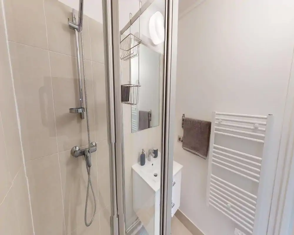 Compact modern bathroom with a tiled shower, glass door, small vanity with sink, mirror and towel radiator. Bright, clean and neatly arranged — good for rental listings.
