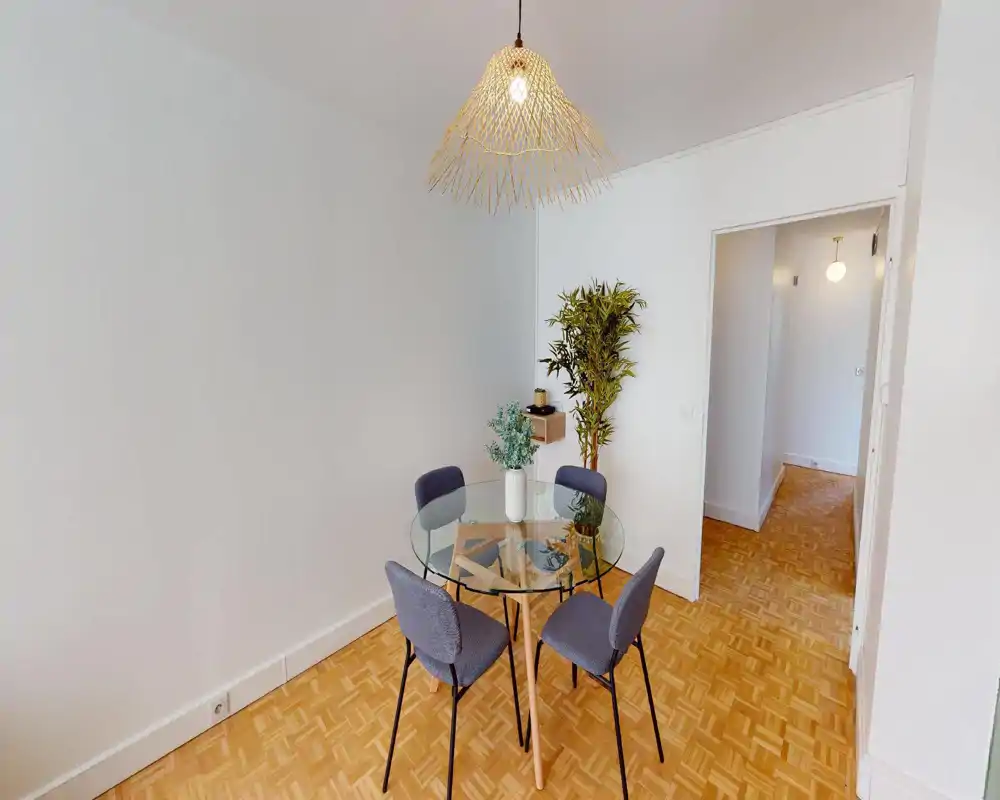 Bright, modern dining area with a round glass table, four upholstered chairs and a decorative plant under a stylish pendant lamp — ideal for small dinners.