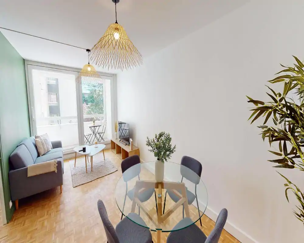 Bright, modern living room with a gray sofa, coffee table and a round glass dining table near a large window opening onto a small balcony. Neutral walls, wooden parquet and decorative plants give a warm, inviting feeling ideal for short-term rentals.