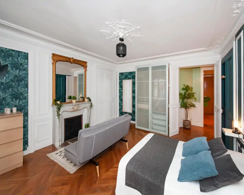 Ampia e elegante camera da letto con parquet, cornici decorative e un comodo letto matrimoniale con biancheria grigia e blu; un divanetto e uno specchio ornamentale aggiungono carattere.