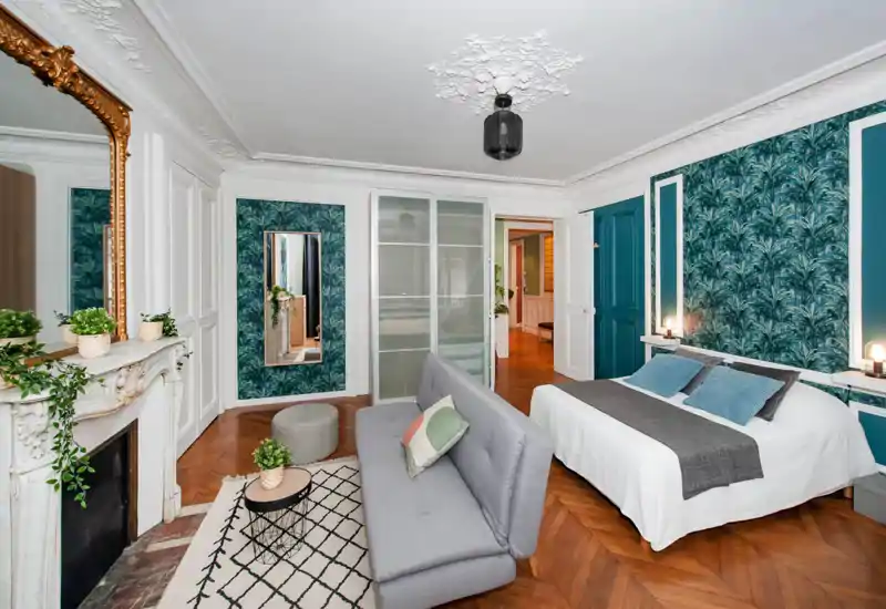 Camera da letto luminosa e di design con un comodo letto matrimoniale, carta da parati turchese decorativa, modanature classiche e un'area soggiorno con divano e tavolino.