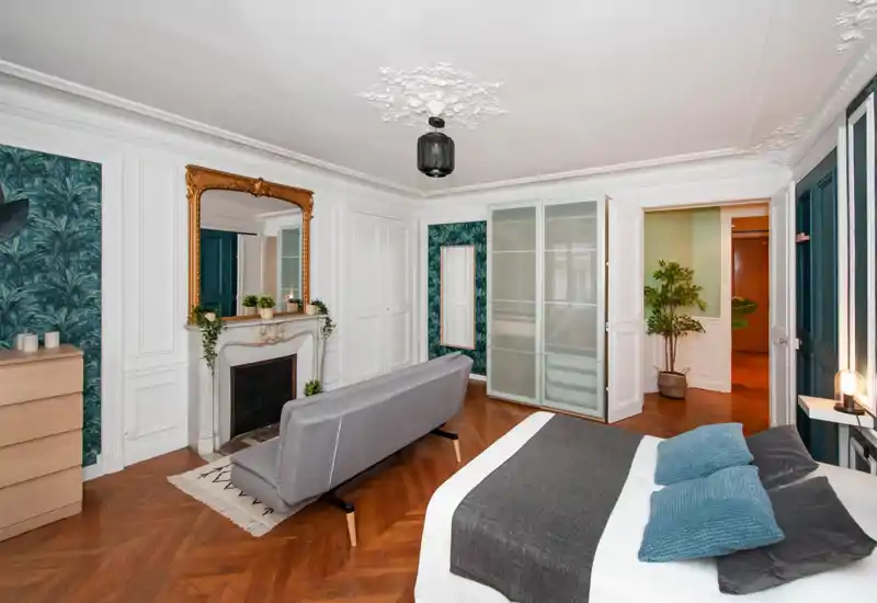 Ampia e elegante camera da letto con parquet, cornici decorative e un comodo letto matrimoniale con biancheria grigia e blu; un divanetto e uno specchio ornamentale aggiungono carattere.