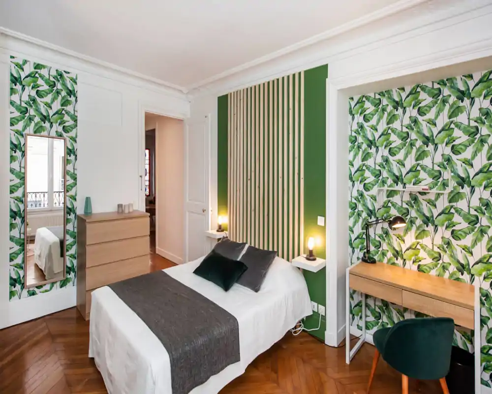 Bright, stylish bedroom with a centrally positioned double bed, leafy accent wallpaper and wooden flooring. The room features bedside lamps, a small desk area and a dresser, offering a cozy, designed stay.