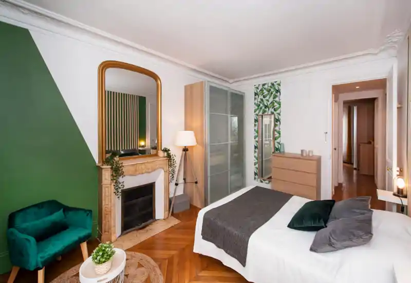 Bright, stylish bedroom featuring a full bed with layered textiles, a decorative fireplace with a large mirror, wardrobe with frosted doors, and leafy accents for a cozy, modern feel.