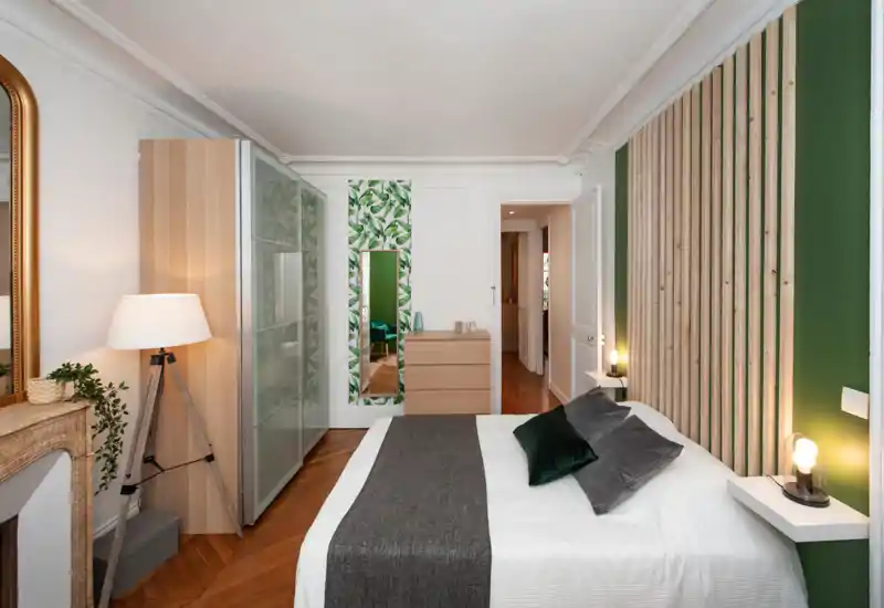 Bright, stylish bedroom with a centrally framed double bed, green accent wall with wooden slats, bedside shelves with lamps and a wardrobe along the left wall. Warm wooden flooring and tasteful decorative details create a welcoming atmosphere ideal for rental listings.