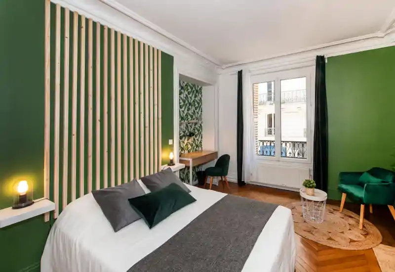 Bright, stylish bedroom with a large bed, green accent wall and natural light from a Parisian-style window. The room features cozy textiles, bedside lamps and a small seating area, ideal for a comfortable stay.